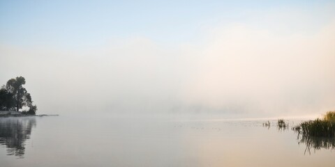 River hidden in a fog. Dense fog over water. Sunrise in mist by the lake on autumn morning. Trees and scírpus-like plants on the bank of the river. © andrbk