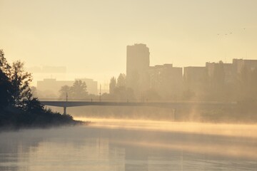 Foggy sunrise in a city. Fog on the water of river canal. Pedestrian bridge and silhouettes of buildings backlit by morning sun. 