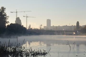 Foggy dawn in a city. Fog on the water of river canal. Bridge and silhouettes of buildings backlit by morning sun. Construction site with cranes nearby.
