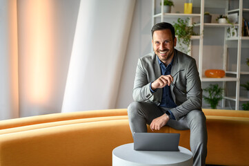 Portrait of handsome business leader holding glasses and smiling while working over laptop in office