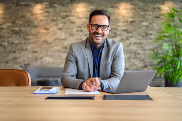 Portrait of handsome smiling entrepreneur with laptop and documents on desk sitting in modern office