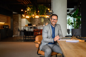 Portrait of happy young professional in eyeglasses with laptop and document on desk working in...
