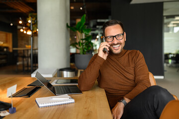 Portrait of cheerful freelancer with laptop and notepad on desk discussing over mobile phone in...