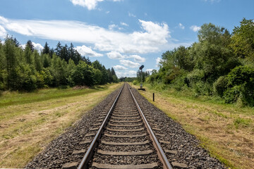 Obraz premium Standard gauge railway tracks with concrete sleepers and a distand light signal in the forest against blue sky with fair weather clouds
