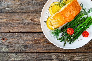 Fried salmon steak with cooked green asparagus, cherry tomatoes and lemon slices served on white plate on  wooden table. Top view