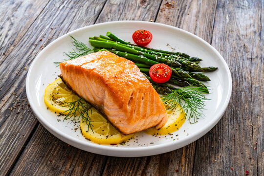 Fried salmon steak with cooked green asparagus, cherry tomatoes and lemon slices served on white plate on  wooden table
