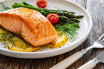 Fried salmon steak with cooked green asparagus, cherry tomatoes and lemon slices served on white plate on  wooden table
