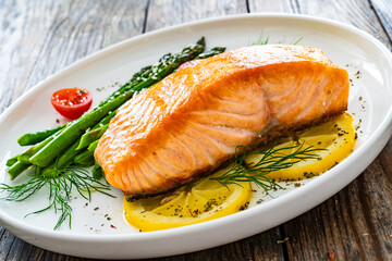 Fried salmon steak with cooked green asparagus, cherry tomatoes and lemon slices served on white plate on  wooden table