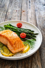 Fried salmon steak with cooked green asparagus, cherry tomatoes and lemon slices served on white plate on  wooden table