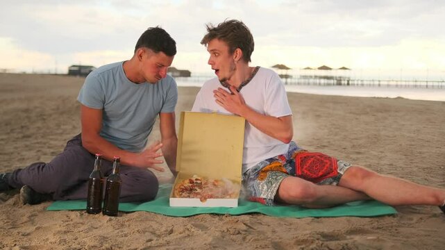 Two cute gay men on a romantic date open the box fresh hot pizza, to enjoy dinner on the beach.
