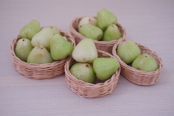 A small wicker basket filled with light green water guava sits on a pale wooden surface. The fruit has a glossy, slightly bumpy texture and a conical shape.