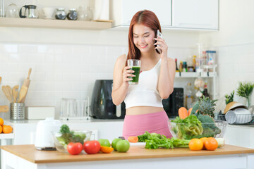 Portrait of a young slim woman talking on phone and drinking fresh vegetable smoothie in the kitchen. Healthy lifestyle concept.