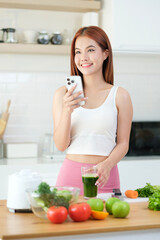 Portrait of beautiful young woman using mobile phone while drinking green vegetable smoothie in the kitchen.