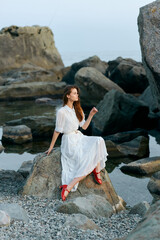 Serene woman in elegant white dress with red shoes sitting on coastal rock, gazing out at sea