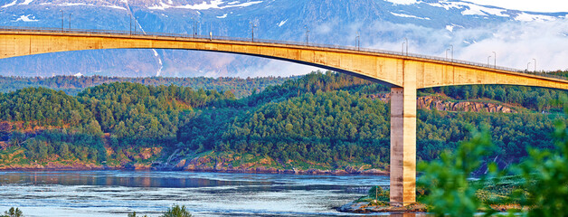 Bridge, road and river with snow on mountains, nature and trees for travel in forest. Landscape, woods and hill with highway, infrastructure and transportation in environment with summer in Norway