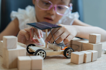 A young girl is building a solar car out of wooden blocks. She is wearing glasses and she is focused on her project