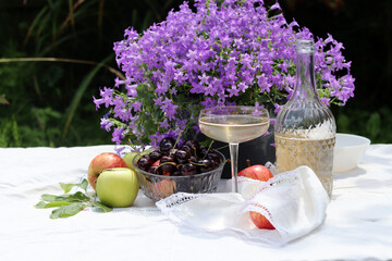 Still life with an open book, cherry, apples, wine glasses and Campanula flowers bouquet on a garden table. Bright picture of picnic table. Summer leisure activity concept. 
