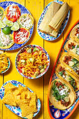 Assortment of colorful Mexican dishes on a vibrant yellow table, featuring tacos, tamales, aztek soup, totopos
