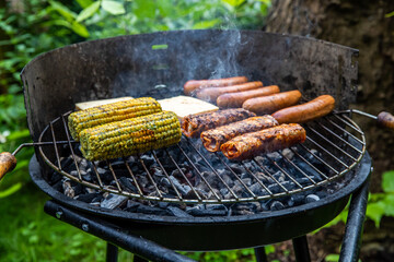 Ein Holzkohlegrill mit Würstchen Maiskolben und Grillkäse belegt.