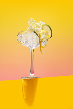 Homemade fresh elderflower lemonade garnished with lemon peel and a blooming branch on a bright pink and yellow background
