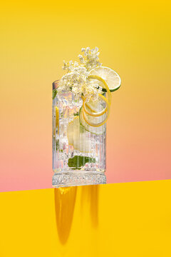 Homemade fresh elderflower lemonade garnished with lemon peel and a blooming branch on a bright pink and yellow background
