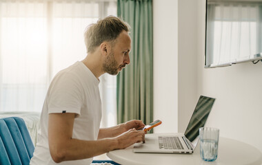 Man working from home using laptop and smartphone, concentrating on tasks, representing modern remote work lifestyle and technology use.