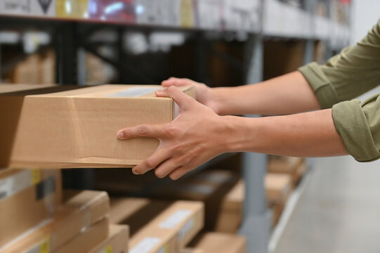 Hand of a person holding a cardboard box in a warehouse, illustrating the act of picking up or storing products. The scene captures a close-up view of the hands.