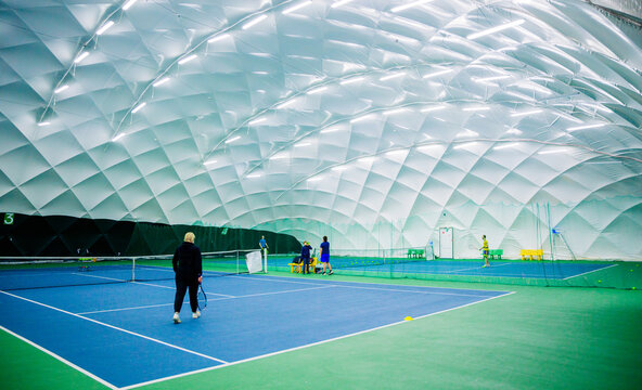 Players practicing on indoor tennis courts. An indoor tennis court with a blue and green surface, featuring multiple players practicing. The court is inside a dome with bright lights.