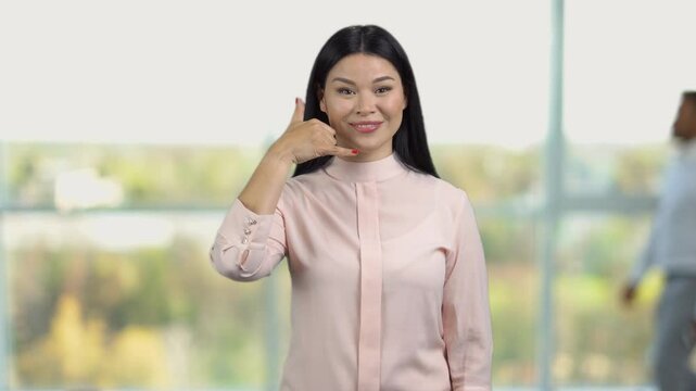 Portrait of a young asian woman shows call me gesture sign. Office window in the background.