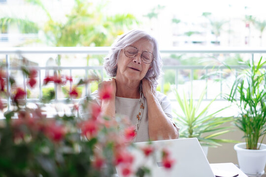 Gray haired senior woman in distress from strong and terrible attack of neck pain after studying on laptop, exhausted senior woman suffering from muscle contracture