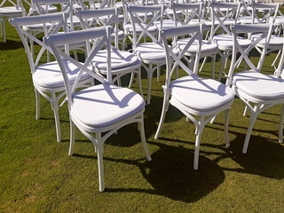 Rows of white folding chairs on lawn before a ceremony. White chairs in rows for public event. Many empty chairs in garden.