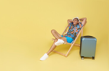 Young asian travel man wear summer clothes sit in deckchair put hands behind head relaxing isolated on yellow background.