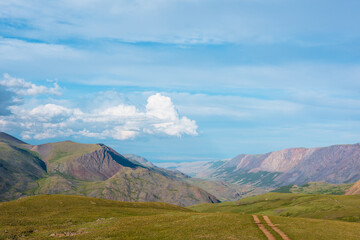 Beautiful top view from grassy green pass with dirt road to wide alpine valley between big mountain range and multicolor mountains under clouds in blue sky. Sunlit large valley in changeable weather.
