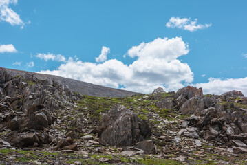 Scenic minimal view to wild mountain pass with green grass and sharp rocks under clouds in blue sky. Minimalist alpine landscape with grassy stony rocky hillside with old outliers in high mountains.