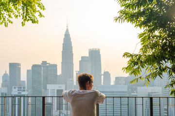 Naklejka premium Back view of man tourist standing on the rooftop terrace of high rise building with green trees and looking at panoramic skyline of Kuala Lumpur city at sunrise, Malaysia. Rear view of male traveler