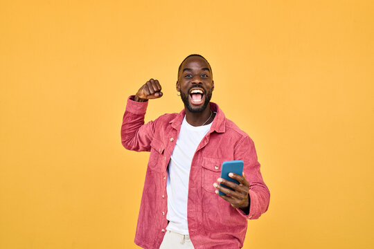 Happy Black man winner holding smartphone celebrating win isolated on yellow. Excited African young gen z guy holding cellphone. Amazed user sport betting game app winner using mobile phone.