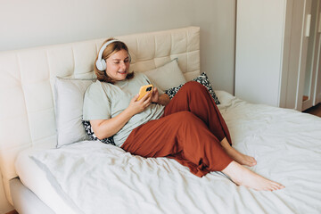 Young 30s woman wear t-shirt and headphones listen music, use mobile cell phone, sits on the bad,...