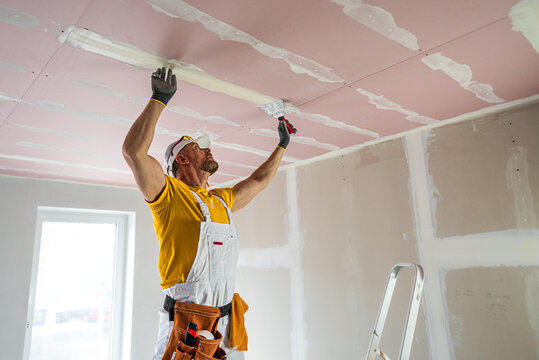 The worker make a plasterboard ceiling. He does taping plaster drywall ceiling joints. A red plasterboard improve the fire resistance of ceiling structures.