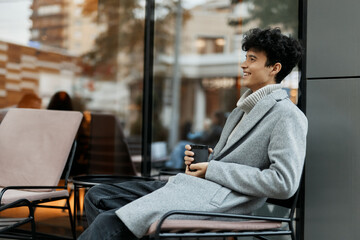 Young man sitting in front of a large window, holding a cup of coffee and smiling