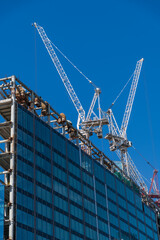 Fototapeta premium Close-up low angle view of group of working cranes on construction site of modern high-rise apartment or office building against blue sky in sunny day. Copy space. Development theme.
