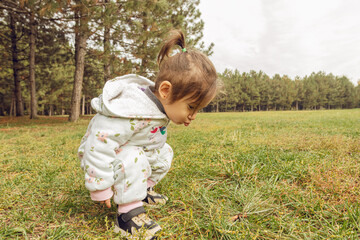 Fototapeta premium Cheerful toddler girl in white floral jumpsuit running in pine forest park. Family lifestyle, childhood development, or outdoor recreation themes.