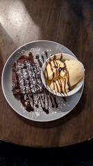 Hot brownie with vanilla ice cream and chocolate sauce on white plate standing on wooden table