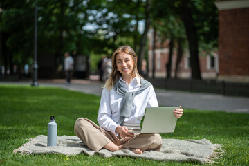 Obraz premium Smiling businesslike woman working remotely online using portable computer. Cheerful female relaxing resting outside outdoors in public place in nature. Joyful woman employee busy with job tasks.