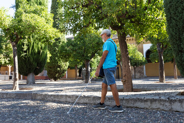 Fototapeta premium A blind mature man walks through a quiet, sunlit park using a white cane. Casual clothes. Peaceful atmosphere. The image captures the essence of independence and resilience.