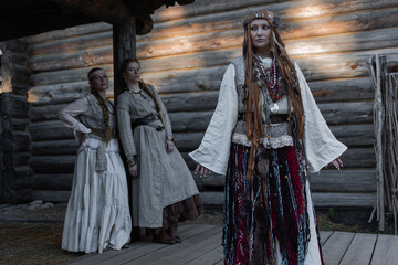 Young girls and a shaman woman in folk costumes, in a village among wooden architecture, Slavic or Nordic style