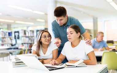 Young male librarian giving advice to positive female students using laptop in the library
