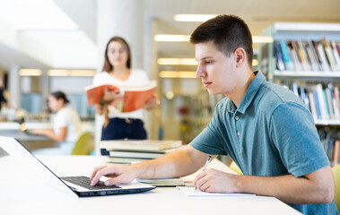 Positive young man in casual clothes working remotely using a computer in a quiet library