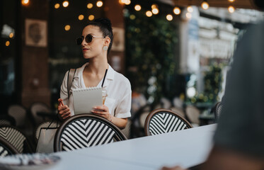 Woman holding a notepad and wearing sunglasses while standing at an outdoor cafe with string lights in the background.