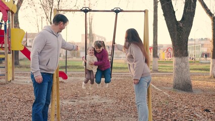 Happy mother father little daughters swing on swing in park autumn. Beautiful little girls sit on swing and laugh. Dad Mom, children play together on playground in fall. Happy family concept, outdoors