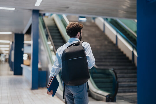 Rear view of a businessman with a backpack and notepad walking through a modern airport terminal towards escalators.